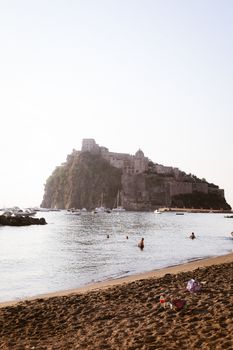 People enjoy a serene beach view at Aragonese Castle, Ischia with boats in the sea.