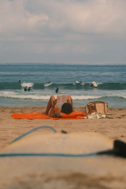 Free Woman reading on a beach while surfers enjoy the waves in the background. Stock Photo