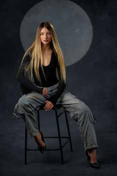 Stylish woman in casual outfit sitting on stool in a dimly lit studio.
