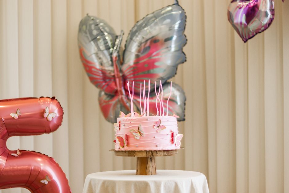 Pink birthday cake with butterfly decorations, surrounded by balloons indoors.
