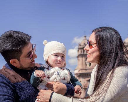 Smiling parents holding their baby in front of a historical building on a sunny day.
