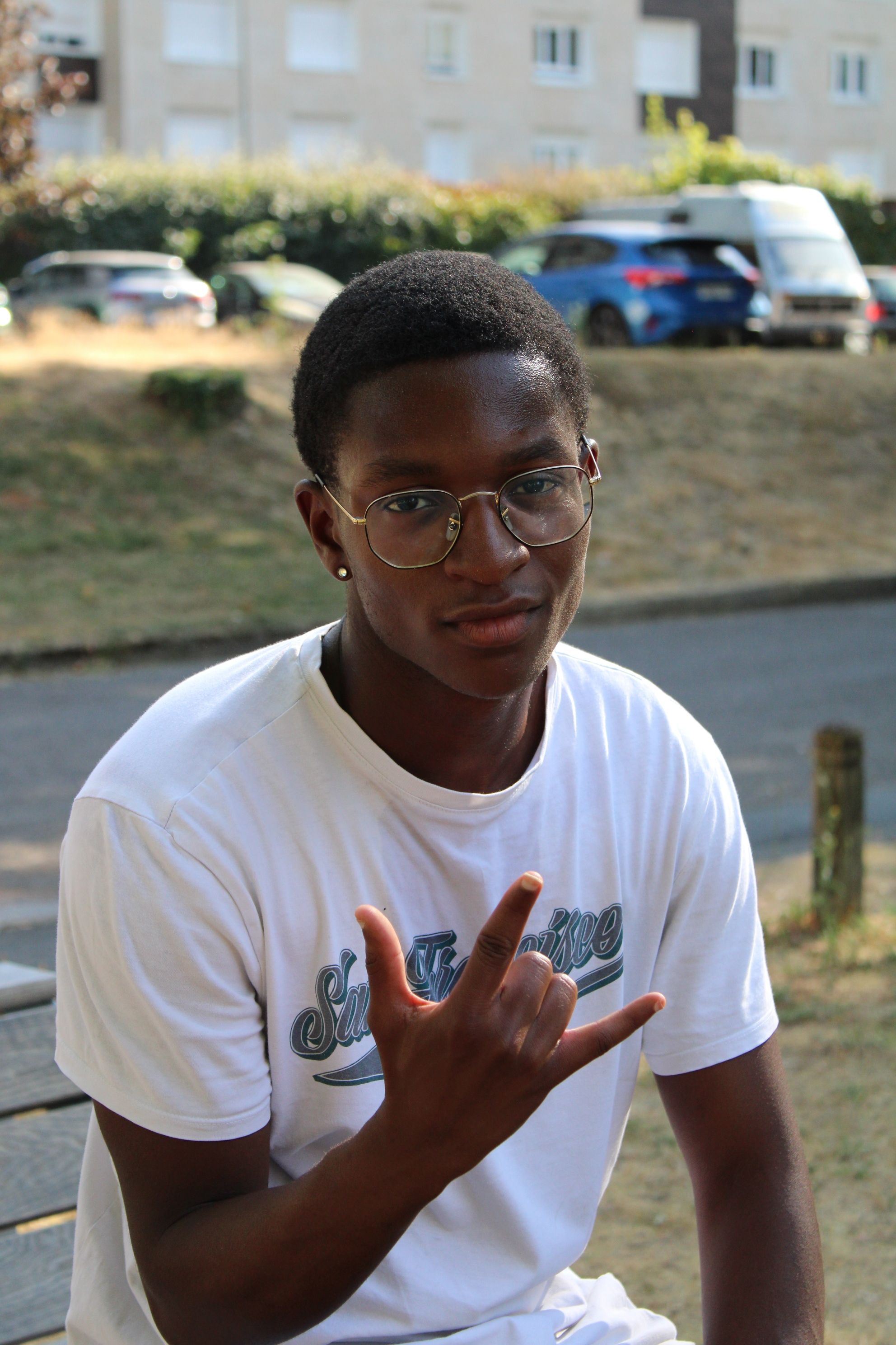 Young man in casual attire making a hand gesture while seated outdoors on a sunny day.