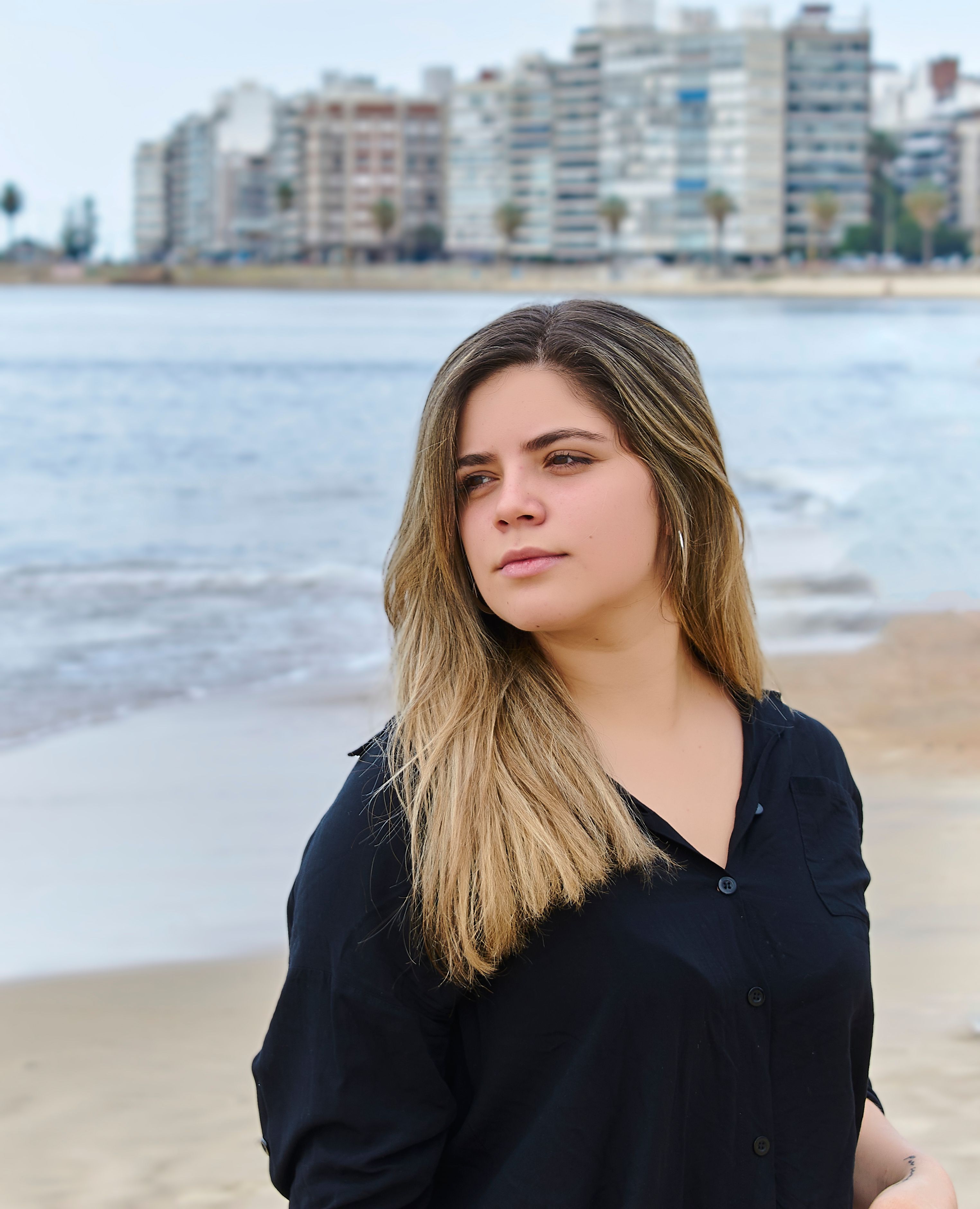 Portrait of a young woman on the beach with Montevideo skyline in the background.