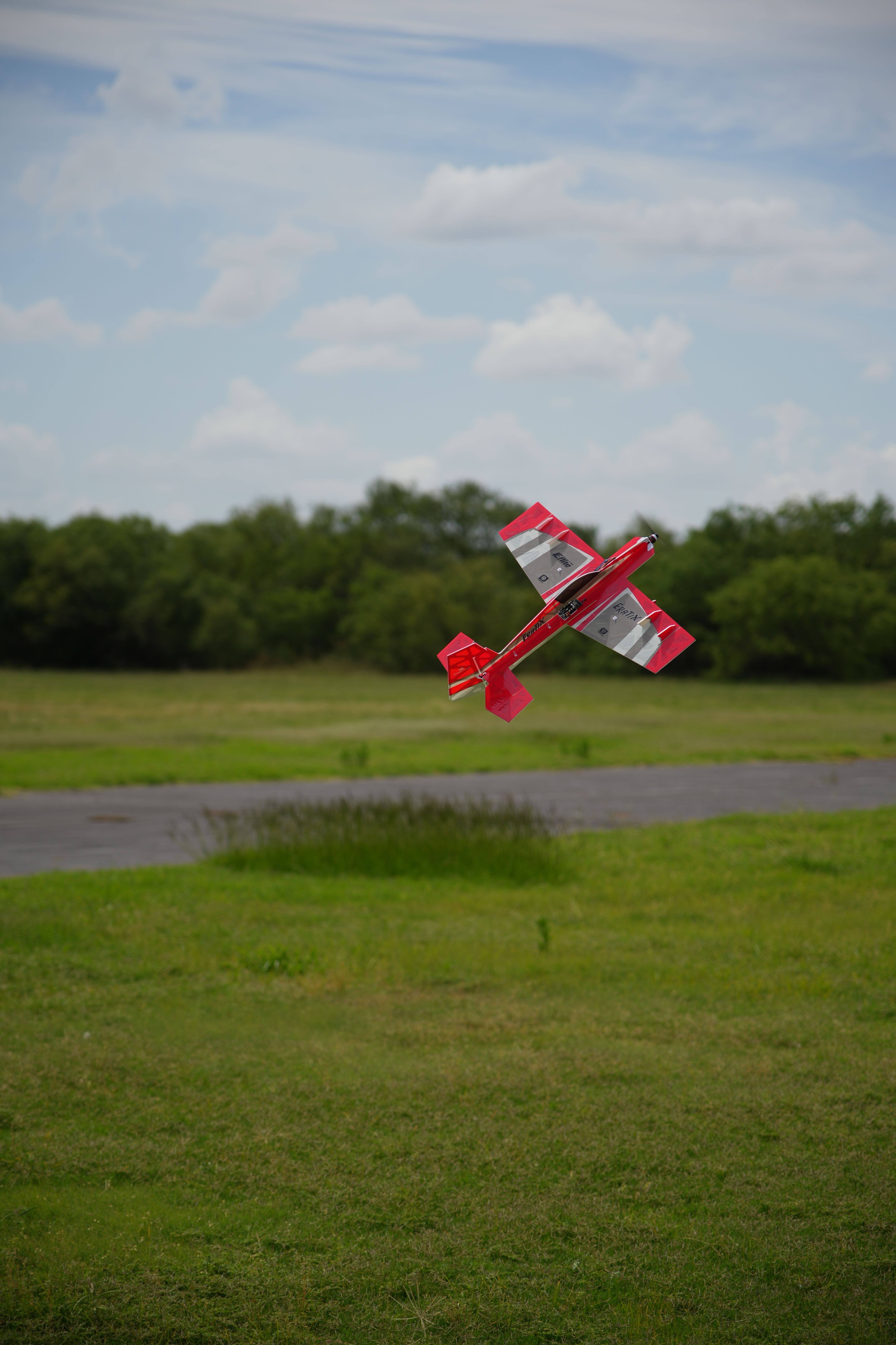 A vibrant red remote-control plane soars over a grassy field with a cloudy sky backdrop, showcasing aerial enjoyment.