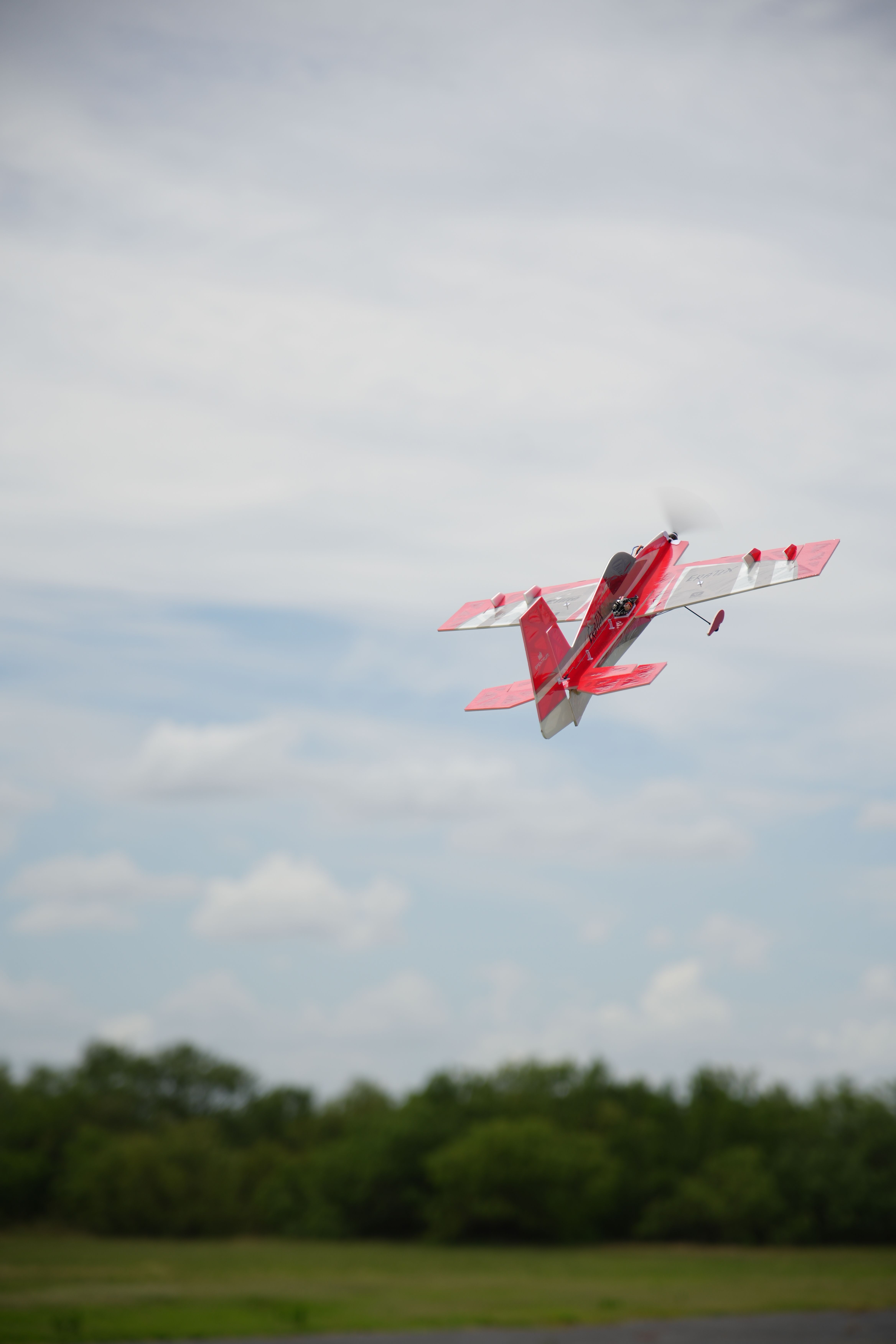 A red remote-controlled plane soaring over a lush green landscape with a cloudy sky.