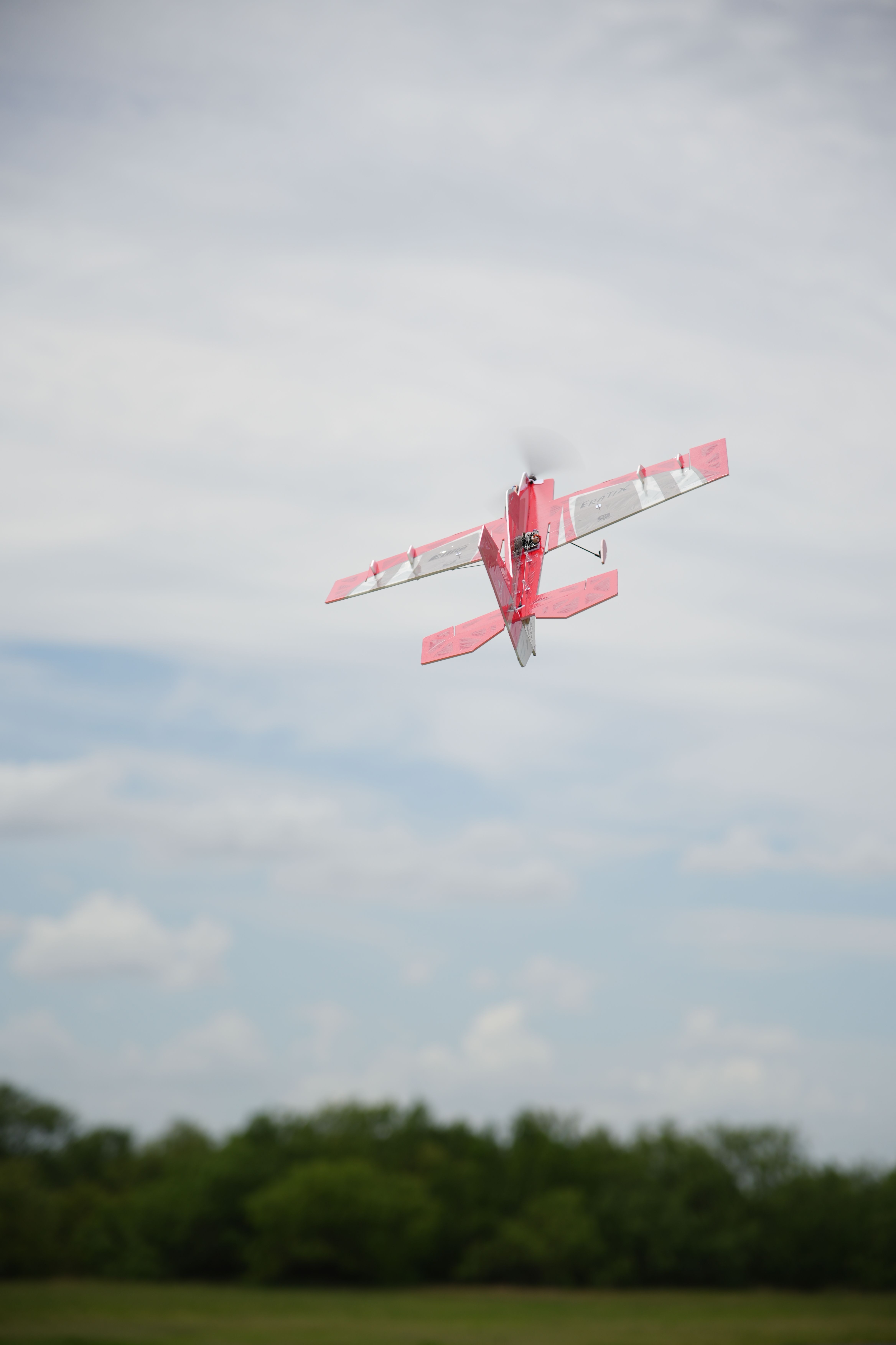 Red RC plane soaring in a cloudy sky over Wichita Falls, Texas, with lush greenery below.
