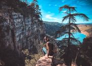 Back view full length male wearing casual clothes sitting on edge of stony cliff against rough mountains and calm blue river while spending time in amazing highlands on clear day