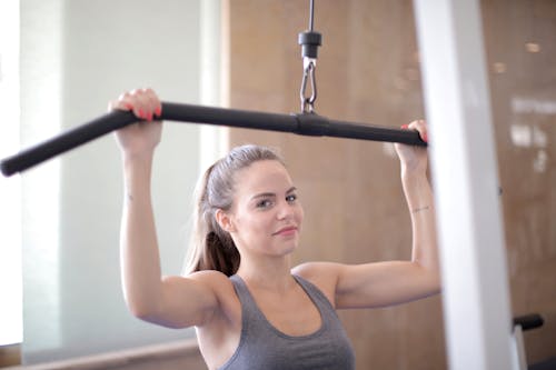 Young friendly muscular woman in sports bra doing exercises using training apparatus and looking at camera during arm training in fitness center