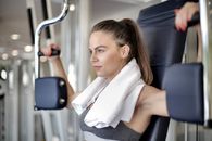 Side view of concentrated powerful female athlete in sportswear and white towel on neck training arm muscles using training apparatus
