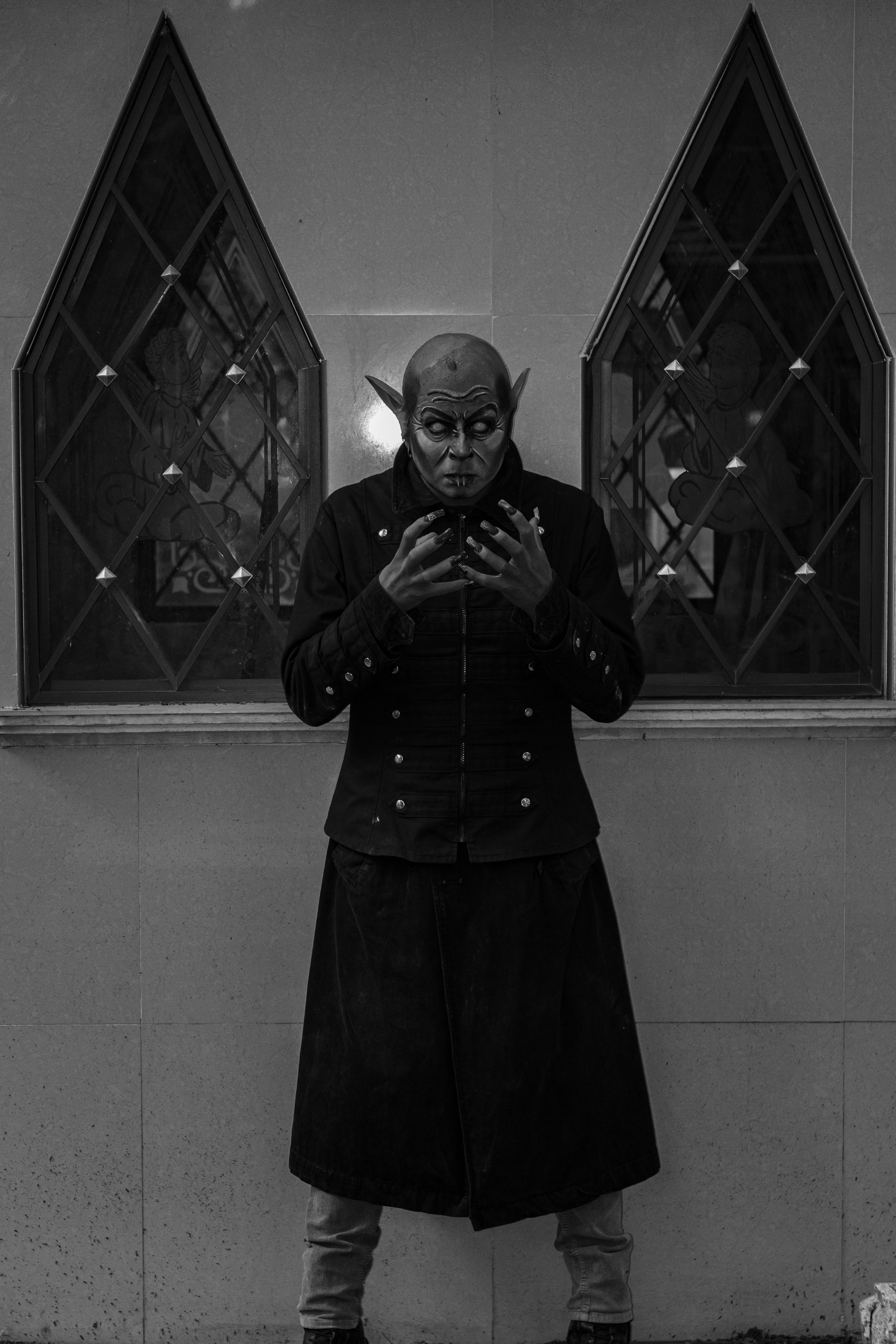 Mysterious figure in dark attire stands before gothic windows, creating an eerie atmosphere.