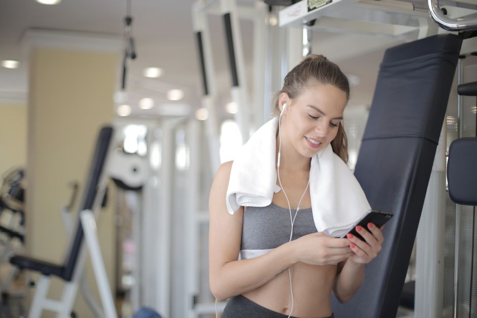 Young smiling lady in gray sports leggings and bra with towel on neck messaging on smartphone and listening to music in earphones during training in gym