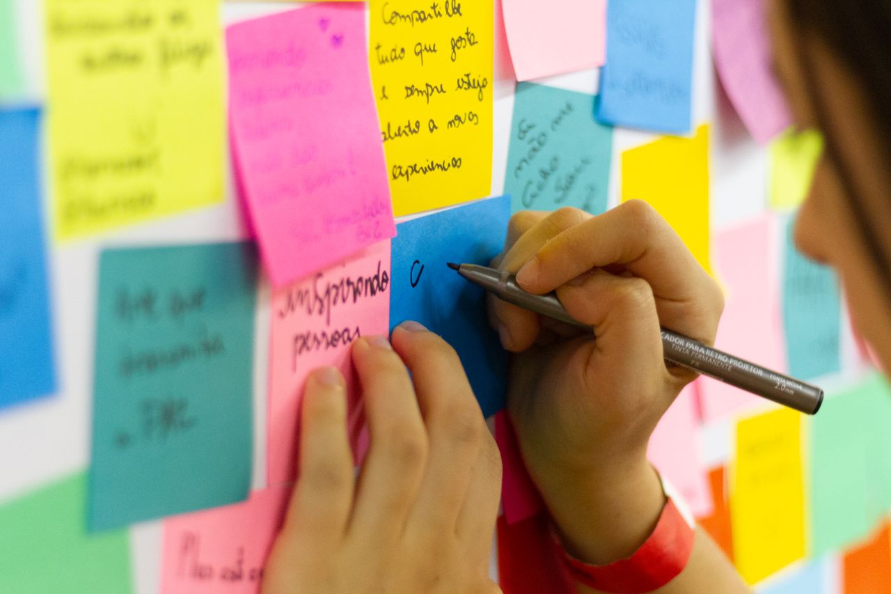 Hands write on colourful sticky notes arranged on a wall.