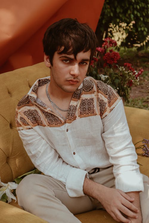 Calm ponder young male in stylish shirt resting on comfy beige chair in summer garden and looking at camera contemplatively