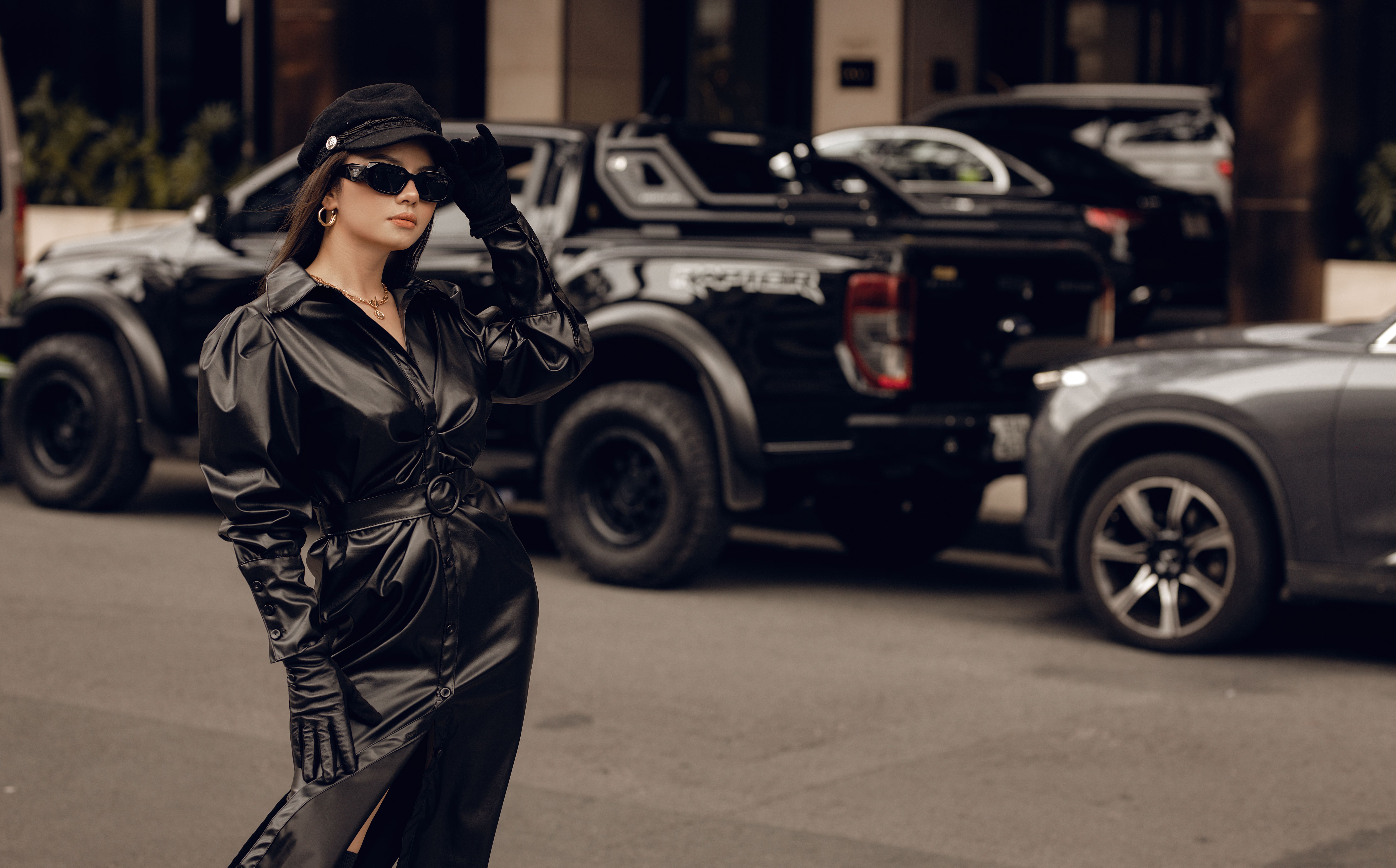Chic woman in black leather dress and sunglasses standing confidently on a city street with vehicles in the background.