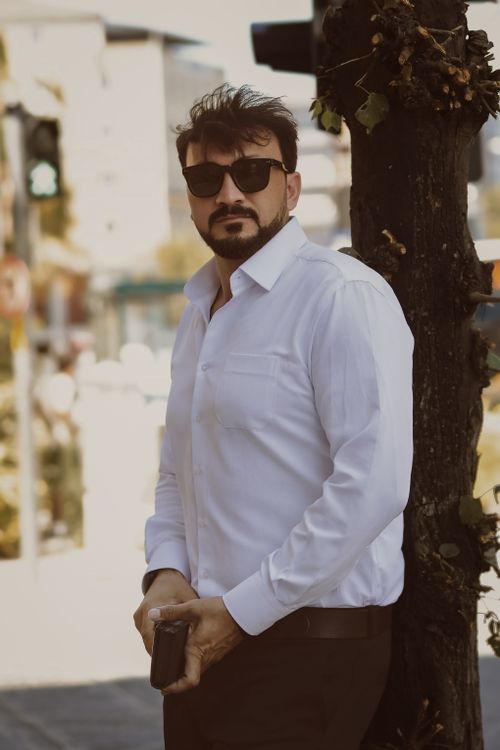 Free Stylish man in white shirt and sunglasses stands against a tree during the day. Stock Photo
