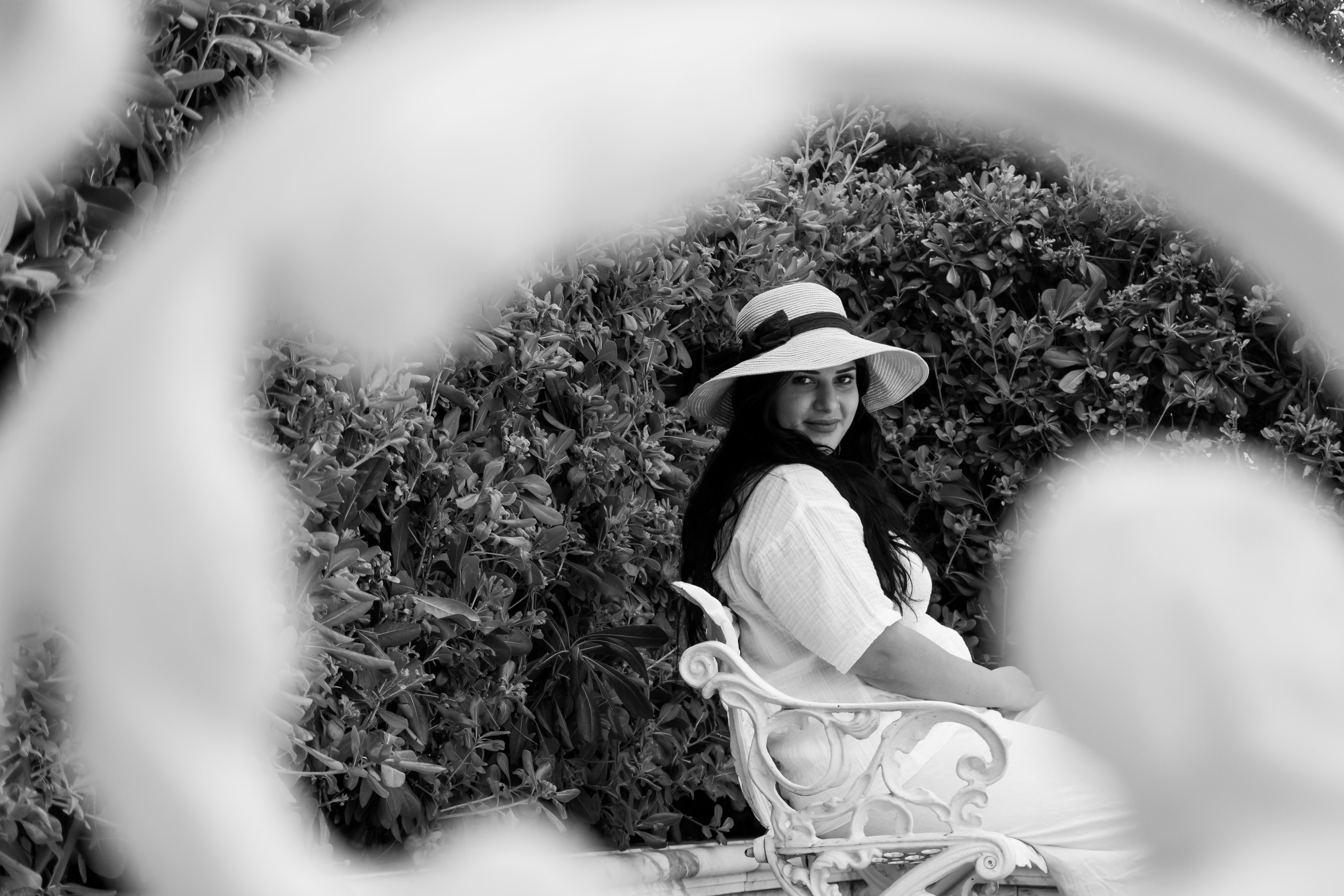 Black and white photo of woman in hat sitting in garden with foliage.