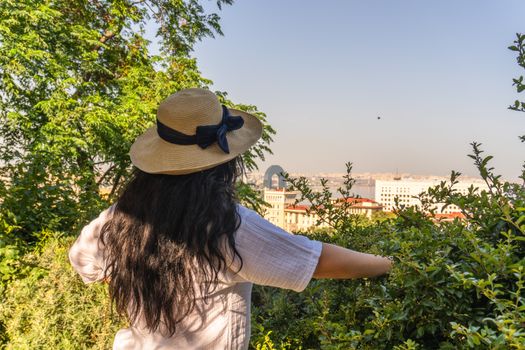 A woman in a straw hat enjoys a scenic view of Baku city from a lush green hilltop.