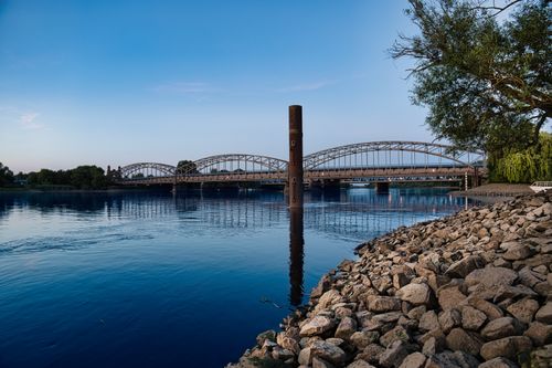 Free Steel bridge over calm river, framed by rocky shore and tree, under a clear blue sky. Stock Photo