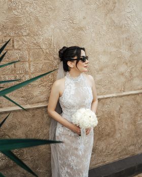 Bride in lace dress wearing sunglasses, holding bouquet against textured wall.