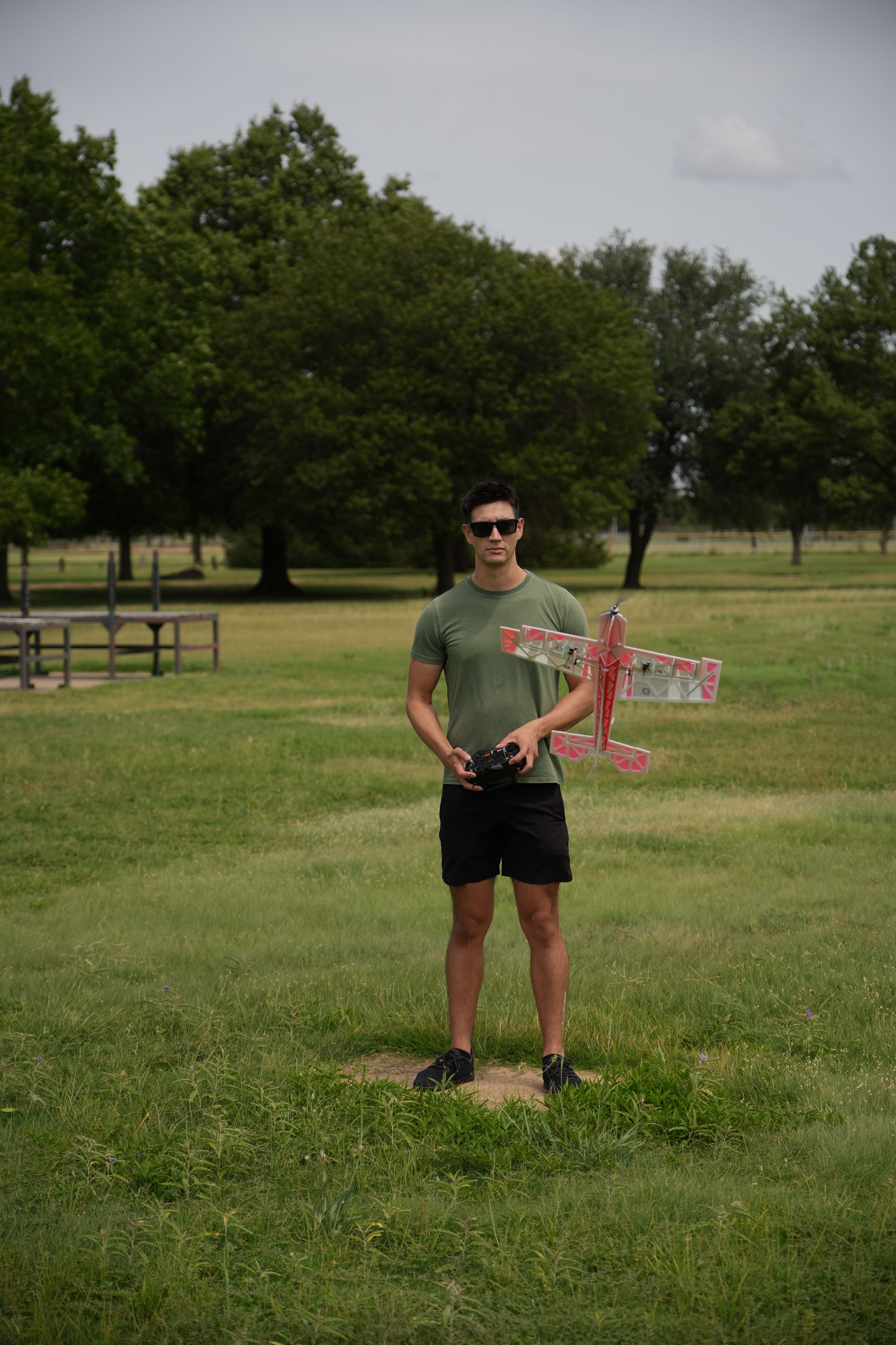 A man operates a remote control airplane in a green park setting under a cloudy sky.