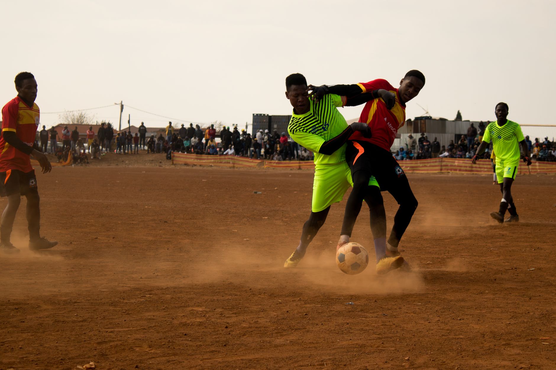 Action-packed scene of a community soccer game with players in vibrant jerseys on a dusty field. — free online jigsaw puzzle on JigsawPuzzlesFreeOnline.org
