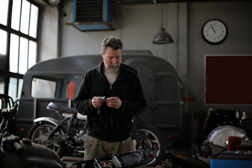 Focused adult bearded male mechanic in workwear standing with details in spacious garage near motorbike and looking down