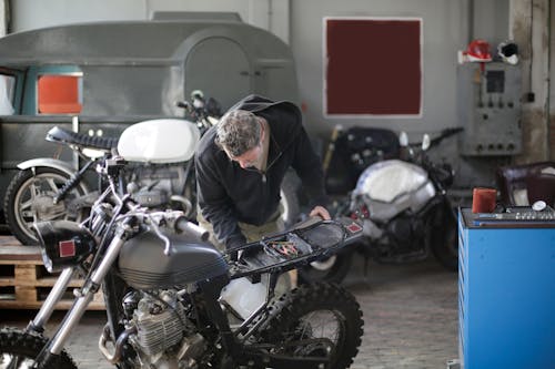 Middle aged bearded male mechanic in workwear and repairing motorbike while standing in modern spacious garage