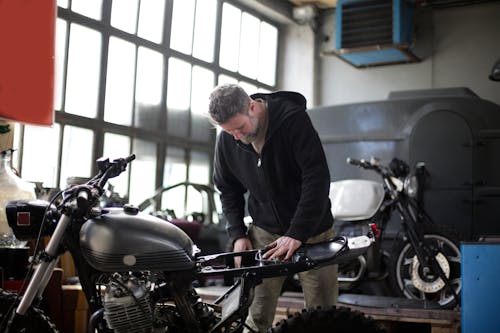 Serious adult bearded male mechanic wearing black workwear standing near motorbike with tools and fixing metal details while working in garage