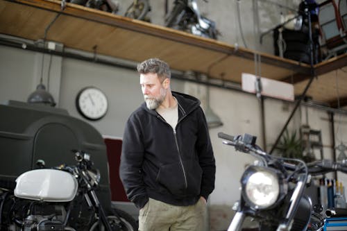 Confident middle aged male mechanic wearing workwear standing with hands in pockets and looking away while repairing motorbike in modern garage