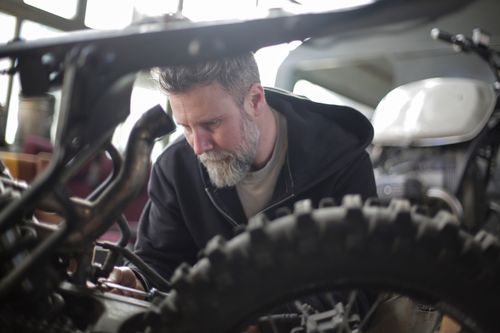 Free Focused adult bearded male mechanic in black workwear sitting near wheel of motorbike with screwdriver while working in modern garage Stock Photo