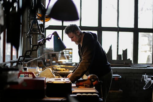Free Side view of serious adult bearded male mechanic in workwear standing near workbench and working with details of engine in light of lamp in garage Stock Photo