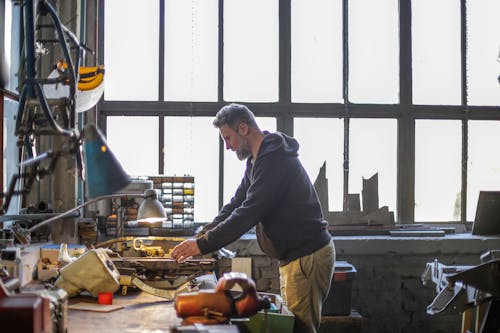 Serious adult bearded worker working with metal details at workbench in workshop