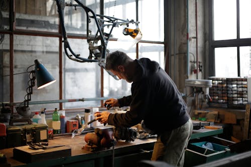Back view of unrecognizable male mechanic in workwear standing near workbench while fixing broken metal detail in lamplight in modern workshop