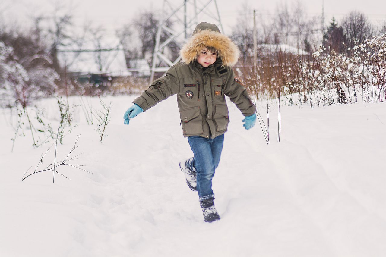 Boy in an olive parka with fur hood, jeans and boots running through deep snow