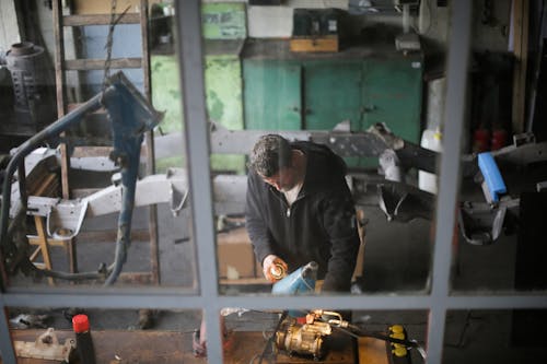 From window view of adult male worker in workwear standing at workbench and fixing metal detail while working in modern garage