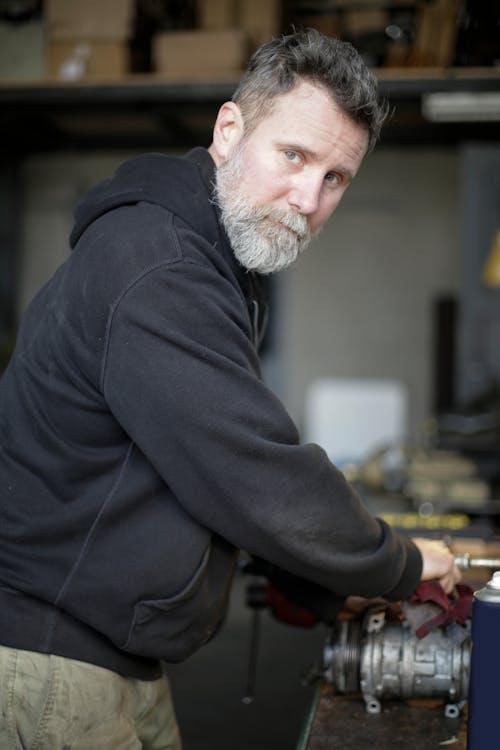 Side view of serious adult bearded mechanic wearing workwear standing near workbench and fixing metal detail of engine while working in garage and looking at camera