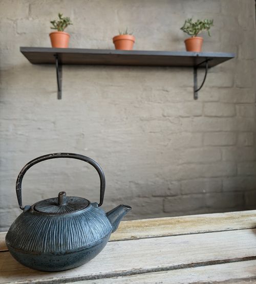 Free A rustic teapot on a wooden table with a shelf of potted plants against a textured wall. Stock Photo