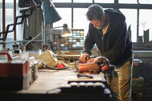 Side view of serious middle aged male in workwear standing near workbench and working with tools and metal details in lamplight in workshop