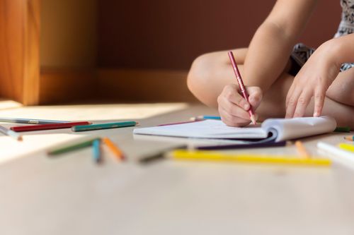 Free Crop anonymous child in casual clothes sitting on floor with legs crossed and drawing in copybook by using multicolored pencils Stock Photo