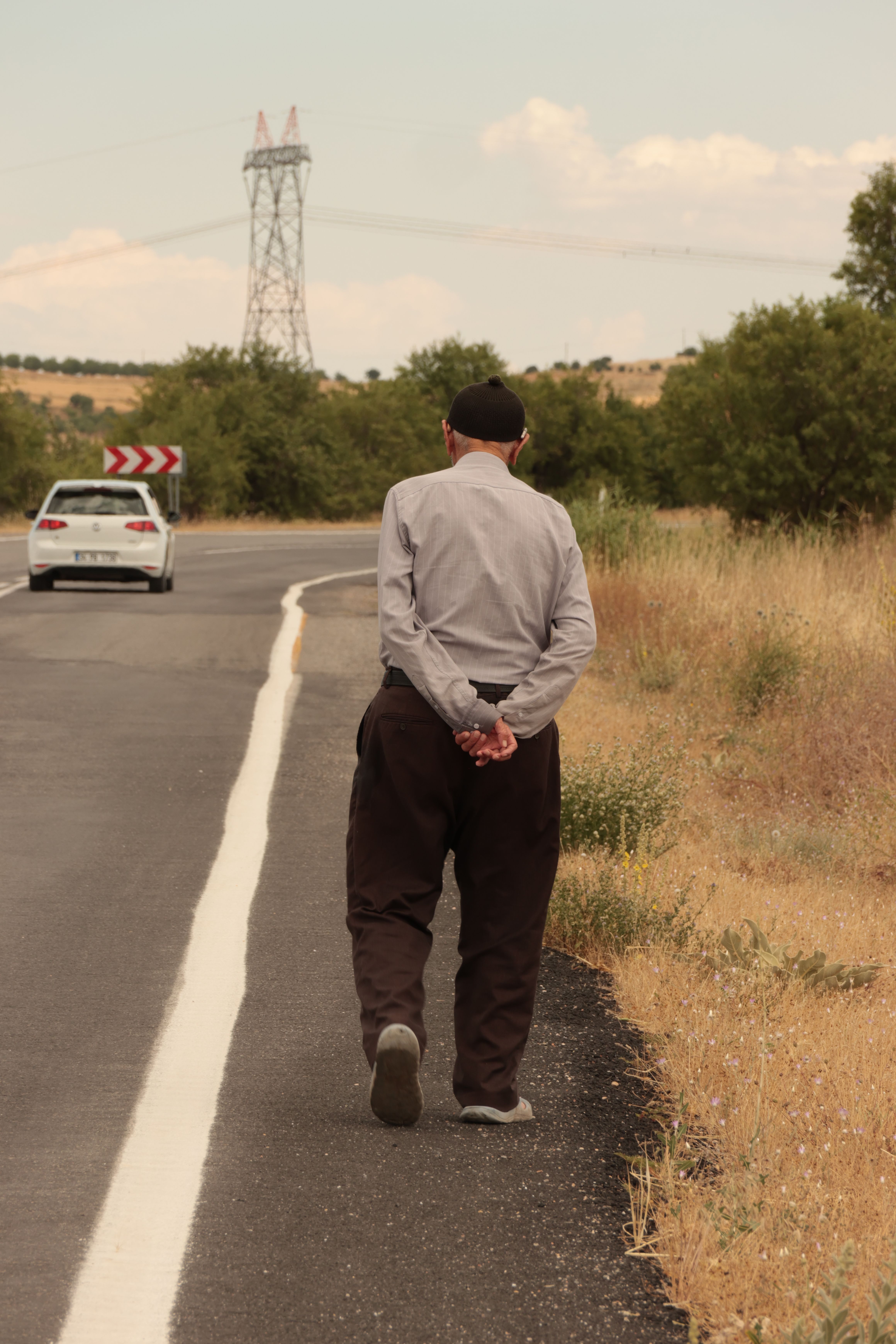 Elderly man strolls down a rural road beside a field on a sunny day