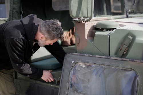 Side view of unrecognizable bearded mechanic in workwear standing near military car and checking details of seat while working on street near garage