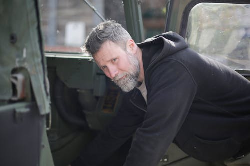 Adult bearded mechanic in workwear standing near military car and examining details while working on street near garage and looking away