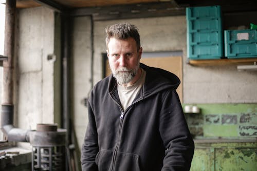 Serious adult bearded mechanic in black workwear standing in spacious workshop and looking at camera