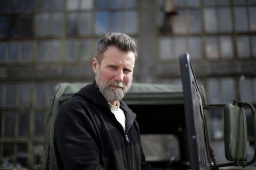 Serious adult bearded man in casual clothes standing near open door of car on street near garage and looking at camera