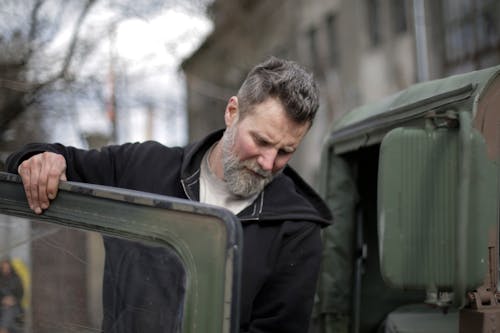 Adult bearded mechanic fixing door of car near garage building