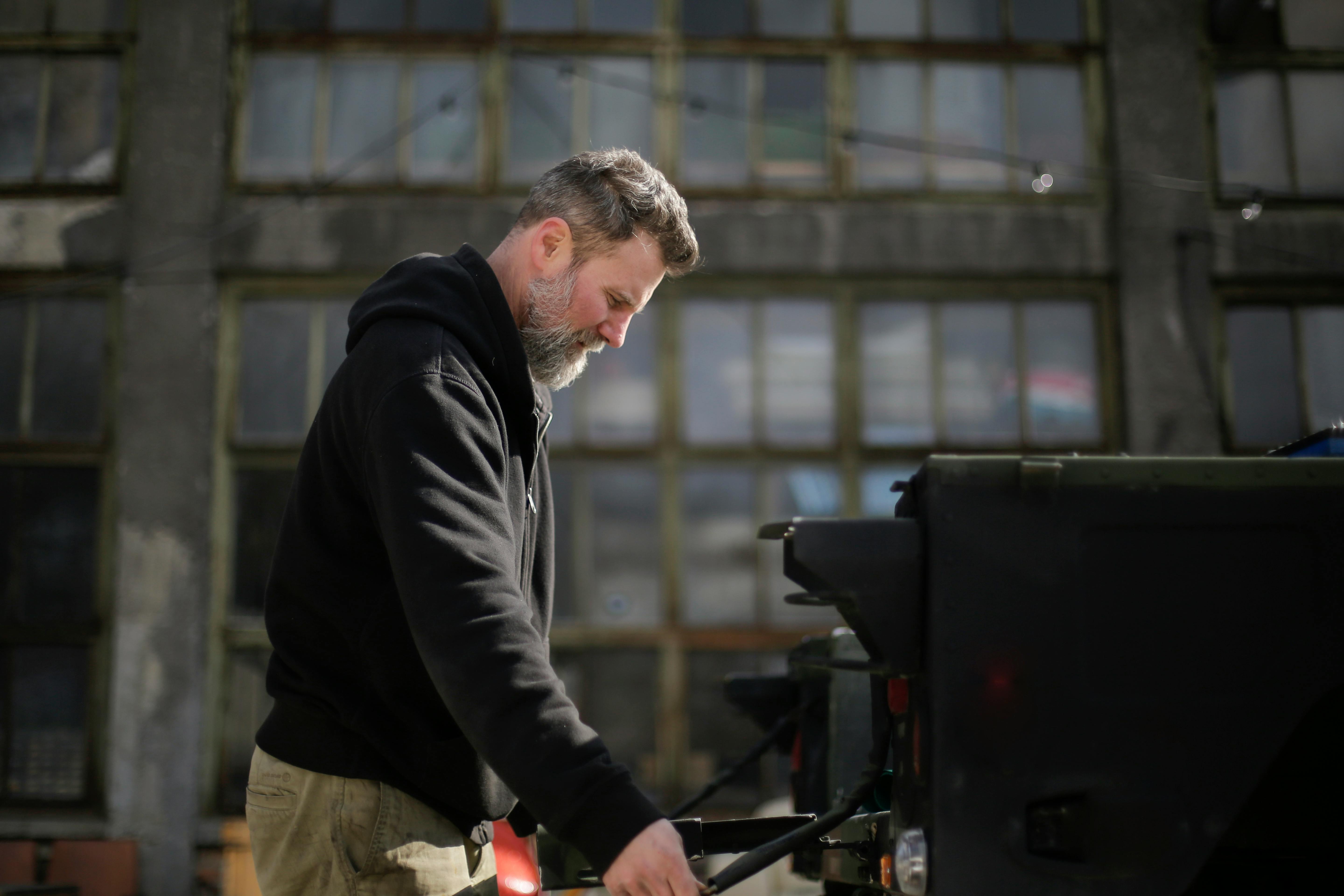 Side view of serious adult bearded mechanic in workwear standing near car and fixing door of trunk on street near garage