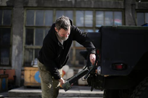 Side view of serious adult bearded mechanic in workwear standing near car on street near garage and repairing metal door of trunk