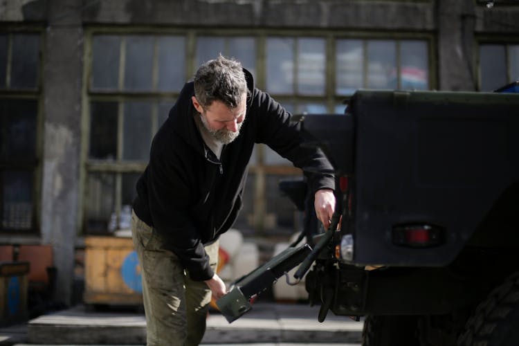 Serious Adult Bearded Mechanic Repairing Metal Door Of Car On Street Near Garage