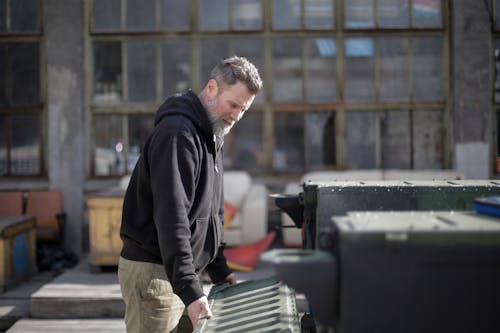 Side view of concentrated adult bearded mechanic in black workwear holding metal door of trunk while examining car on street near garage