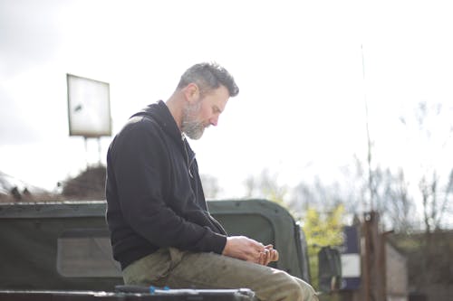 Serious bearded man sitting on metal platform on car outside garage